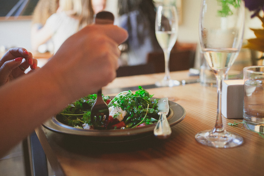 A person uses a fork to eat a green salad at a wooden table with a glass of white wine and water nearby. Jessica Hill Photography - Wedding Photographer - Oregon