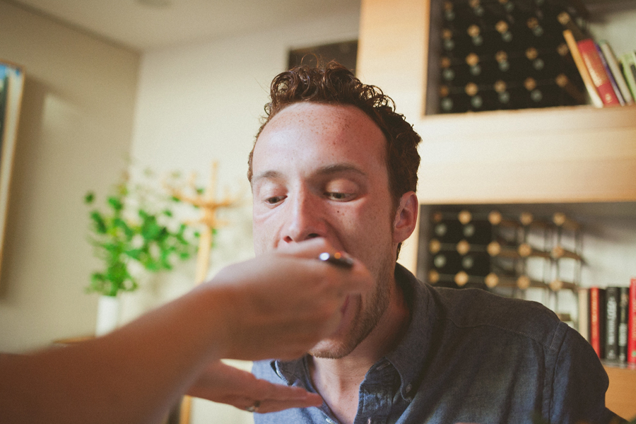 A man is being fed with a spoon by someone else in a home setting, with shelves and a wine rack visible in the background. Jessica Hill Photography - Wedding Photographer - Oregon