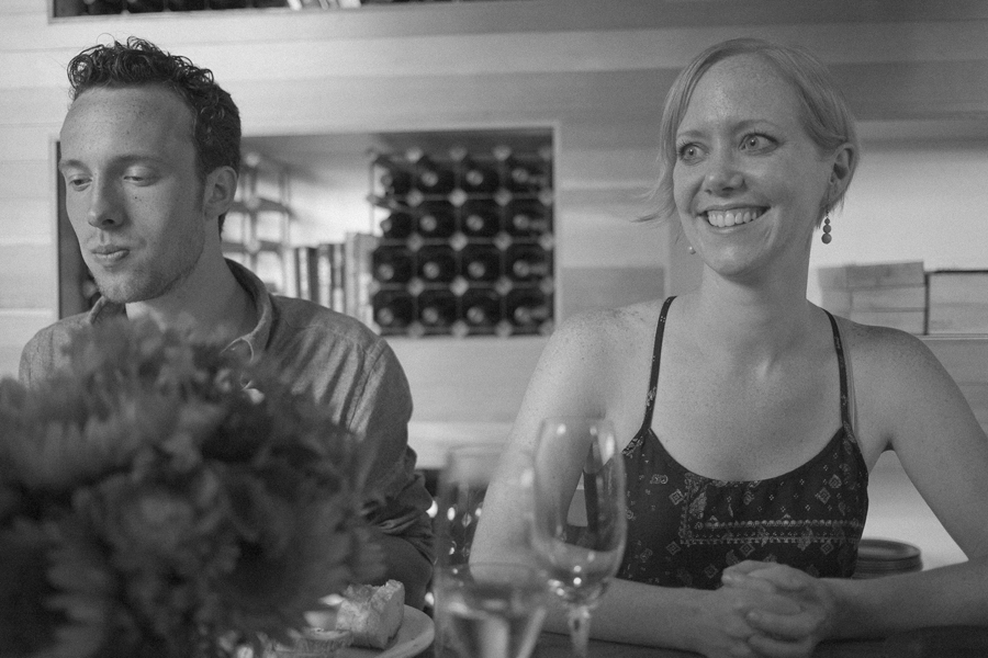 Two people sit at a dining table in a restaurant. The man looks down with a neutral expression, while the woman smiles. Wine glasses and flowers are on the table. Jessica Hill Photography - Wedding Photographer - Oregon
