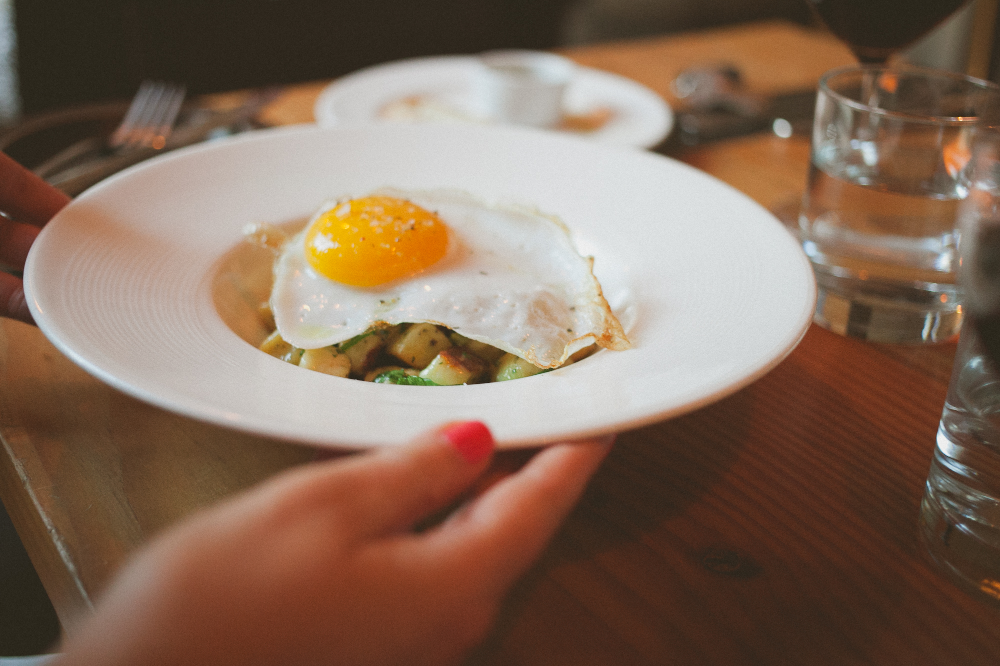 A person serves a white plate with a sunny-side-up egg on top of diced vegetables on a wooden table set with utensils, a water glass, and a glass of red wine. Jessica Hill Photography - Wedding Photographer - Oregon