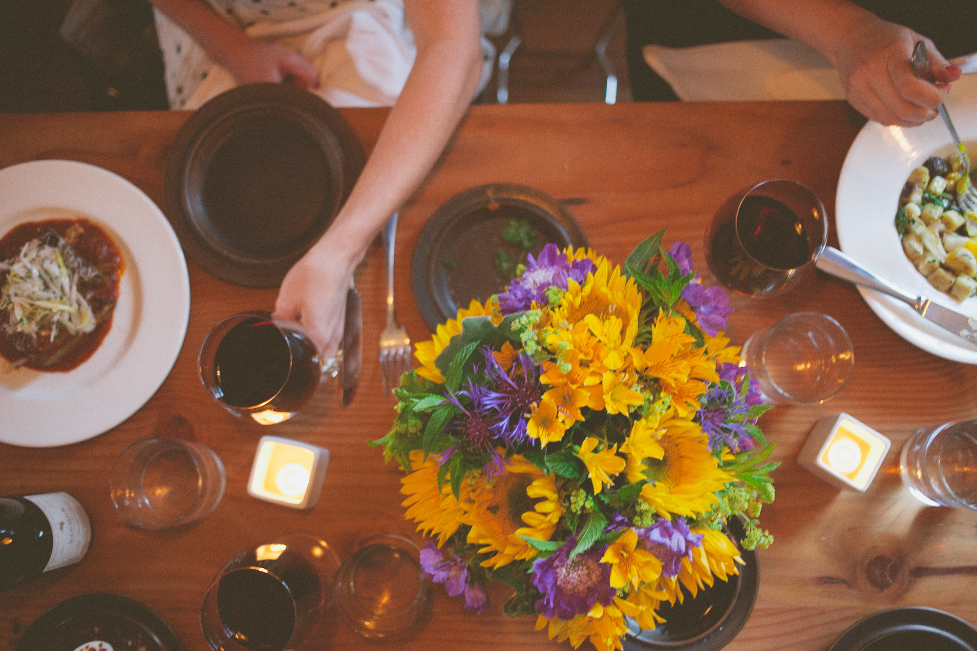 Overhead view of a wooden dining table set for a meal, featuring plates of food, wine glasses, a vibrant bouquet of flowers, and two people seated. Jessica Hill Photography - Wedding Photographer - Oregon