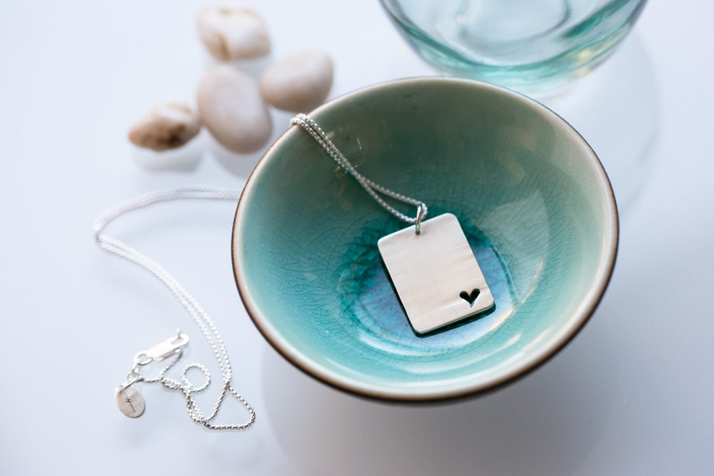 A silver necklace with a rectangular pendant featuring a small heart is displayed in a turquoise ceramic bowl, with smooth white stones and a glass dish in the background. Jessica Hill Photography - Wedding Photographer - Oregon
