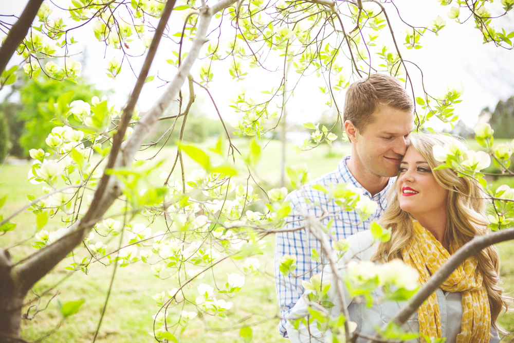 A man and woman stand closely together outdoors among budding tree branches, both smiling and facing slightly toward each other. Jessica Hill Photography - Wedding Photographer - Oregon