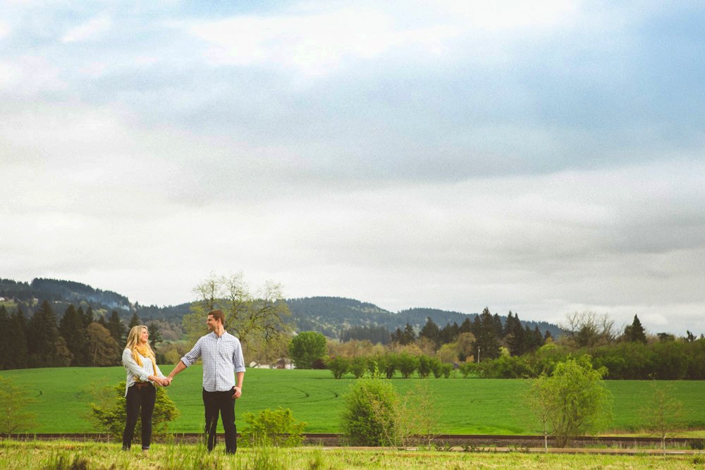 A man and woman stand holding hands in a grassy field with trees and hills in the background under a cloudy sky. Jessica Hill Photography - Wedding Photographer - Oregon