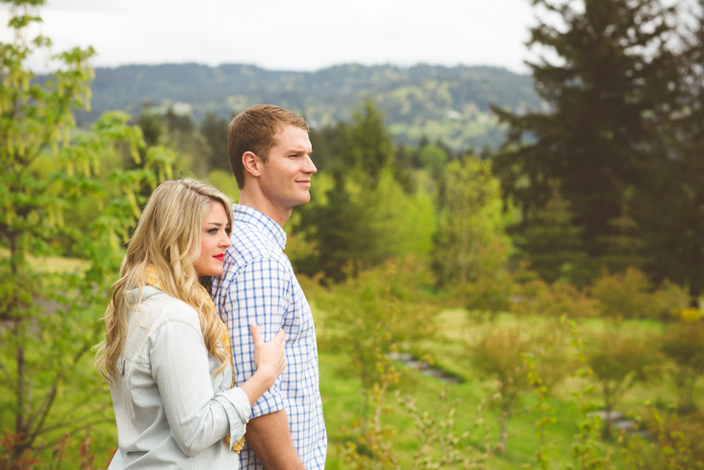 A woman stands behind a man outdoors in a green, wooded area, resting her hand on his arm. Both look toward the distance. Jessica Hill Photography - Wedding Photographer - Oregon