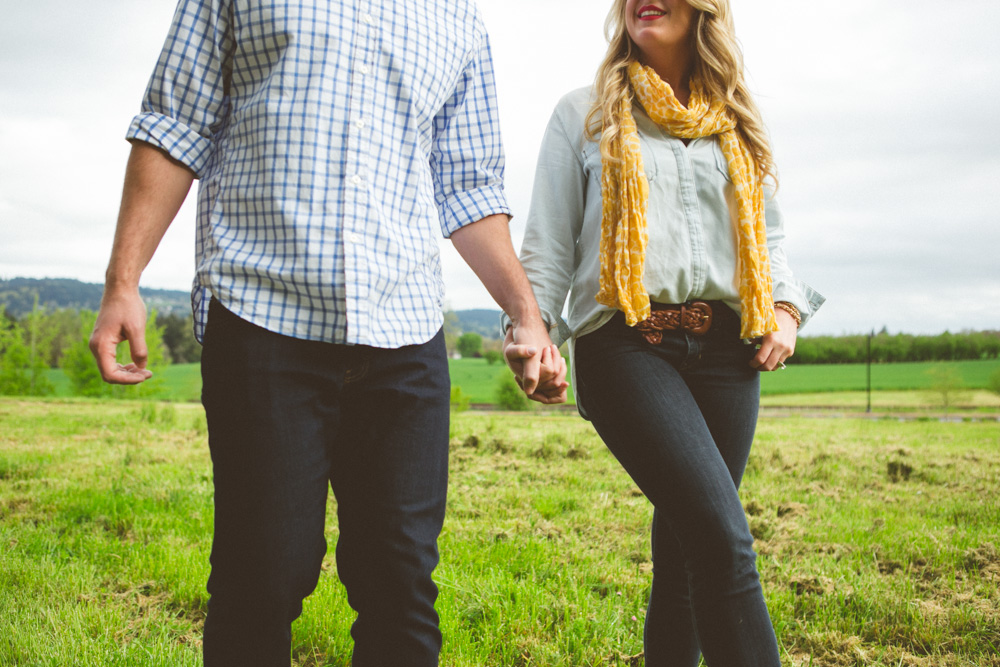 A man and woman holding hands, standing in a grassy field. Their faces are partially out of frame, and both are casually dressed. Jessica Hill Photography - Wedding Photographer - Oregon