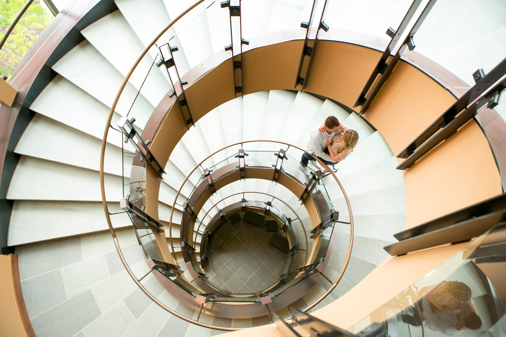 Top-down view of a spiral staircase with two people standing on a landing, surrounded by glass and metal railings. Jessica Hill Photography - Wedding Photographer - Oregon