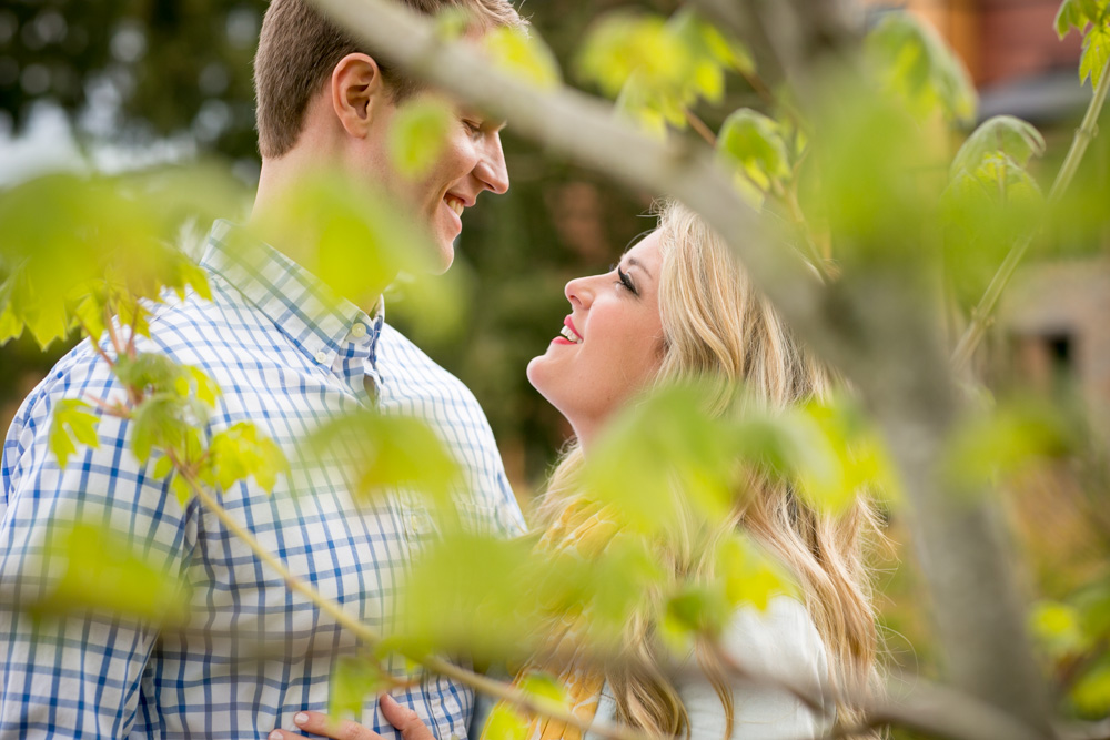 A man and woman stand close together outdoors, smiling at each other, partially obscured by green leaves in the foreground. Jessica Hill Photography - Wedding Photographer - Oregon