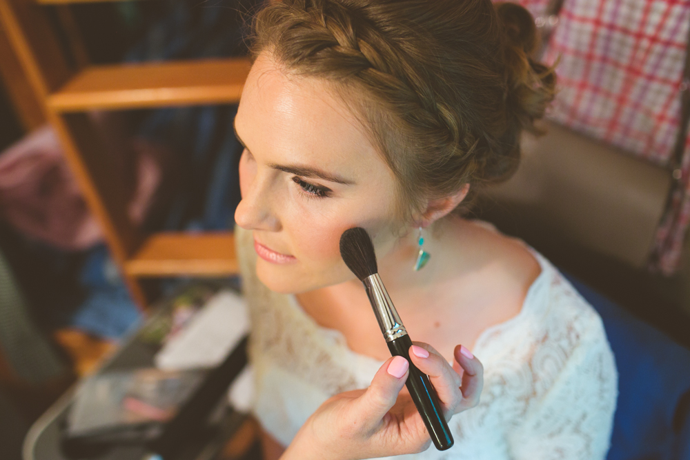 A woman in a white top is having makeup applied to her face with a brush. She has braided hair and is looking slightly to the side. Jessica Hill Photography - Wedding Photographer - Oregon