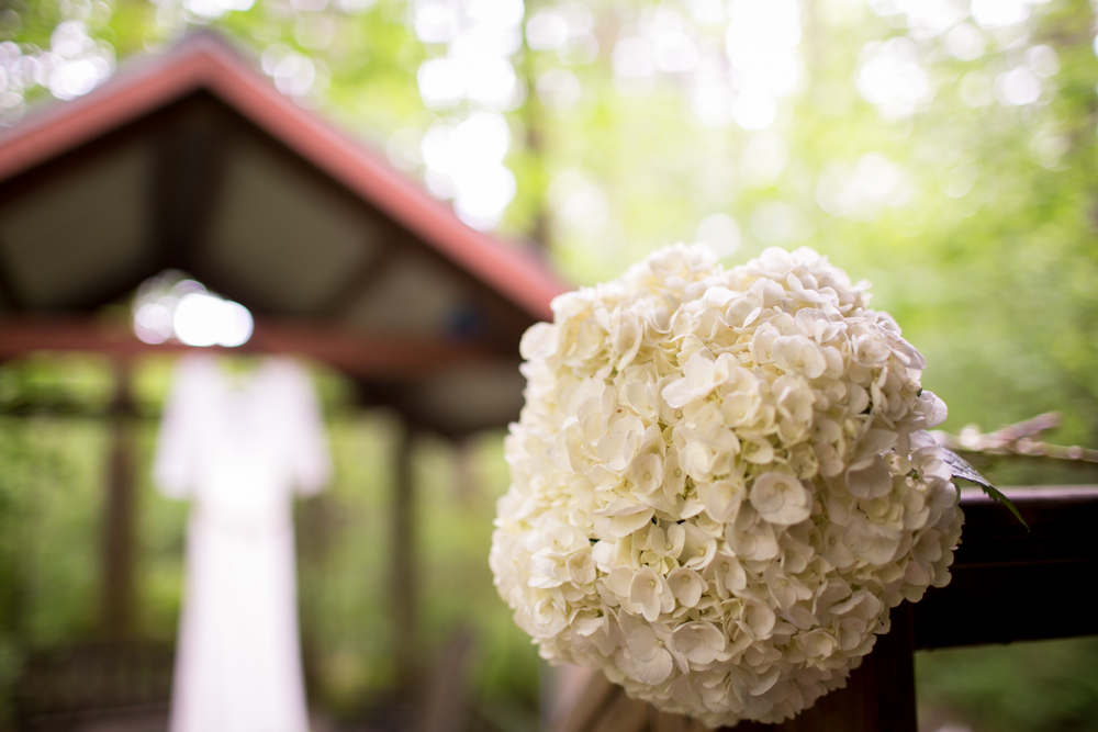 A close-up of a white hydrangea bouquet on a wooden railing with a blurred gazebo and white dress in the background. Jessica Hill Photography - Wedding Photographer - Oregon