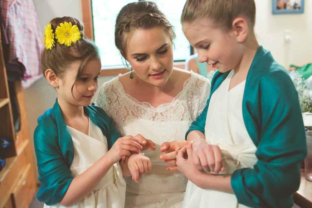 A bride in a white dress sits with two young girls in white dresses and teal shawls, all looking at something on their hands. Jessica Hill Photography - Wedding Photographer - Oregon