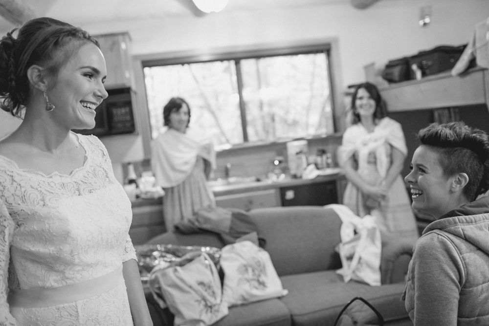 Four women are gathered in a cozy room, with two of them smiling at each other in the foreground. The scene appears casual, with bags and belongings visible around the room. Jessica Hill Photography - Wedding Photographer - Oregon