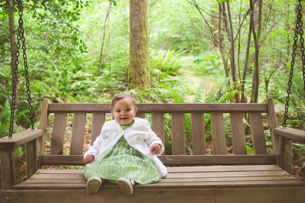 A young child in a green dress and white sweater sits on a wooden swing bench in a lush, green forest setting. Jessica Hill Photography - Wedding Photographer - Oregon