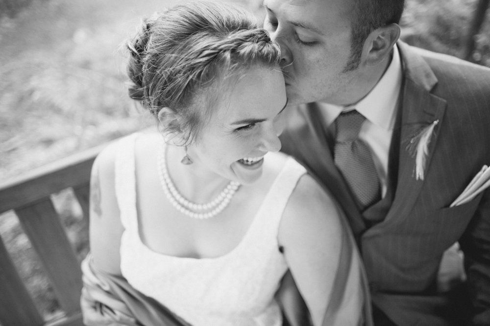 A groom in a suit kisses a smiling bride on the forehead as they sit on a bench; the photo is in black and white. Jessica Hill Photography - Wedding Photographer - Oregon