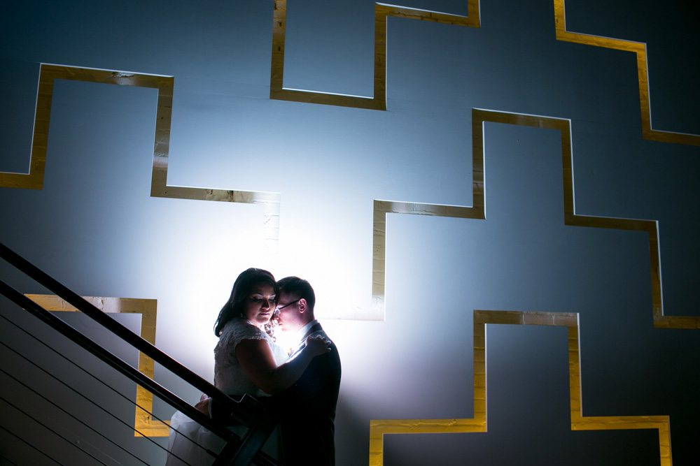 A couple stands closely on a staircase, dramatically backlit against a geometric wall with gold-lined shapes, creating a romantic and artistic silhouette. Jessica Hill Photography - Wedding Photographer - Oregon