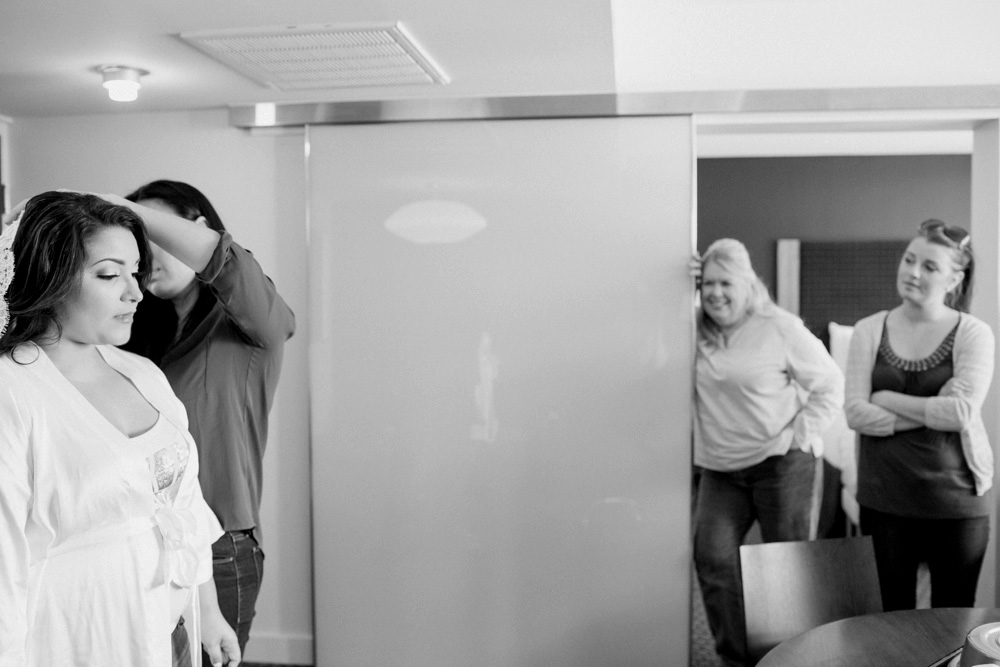 A bride gets her hair styled by a woman, while two women watch and smile from the doorway in a softly lit, modern room. The scene is candid and relaxed. Jessica Hill Photography - Wedding Photographer - Oregon