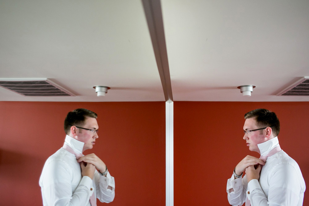 A man wearing glasses adjusts his shirt collar while looking at his reflection in a mirror, with a red wall background and ceiling lights above. Jessica Hill Photography - Wedding Photographer - Oregon