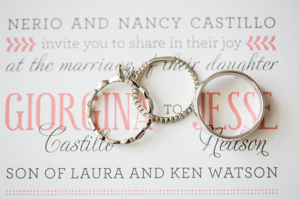 Three wedding rings rest on top of a wedding invitation featuring the names "Giorgina" and "Jesse" in large pink and gray text, along with additional family names and details in elegant fonts. Jessica Hill Photography - Wedding Photographer - Oregon
