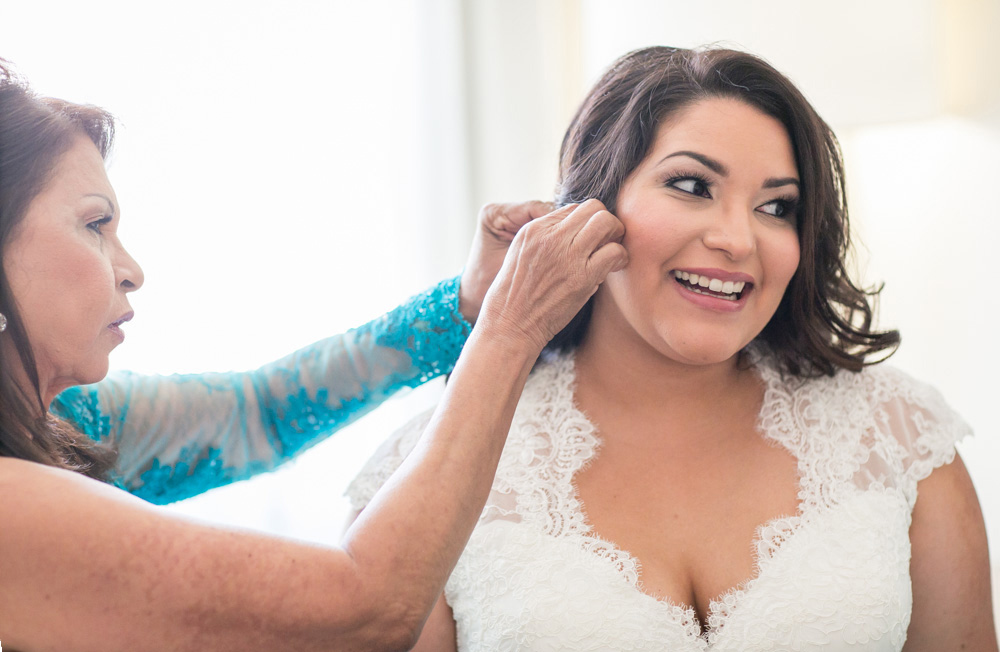 A woman in a white lace wedding dress smiles as another woman in a blue lace sleeve helps her put on earrings, preparing for the wedding. Jessica Hill Photography - Wedding Photographer - Oregon