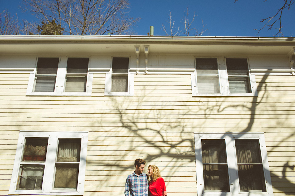 A man and woman stand close together in front of a beige two-story house with several windows and tree branch shadows on the wall. Jessica Hill Photography - Wedding Photographer - Oregon