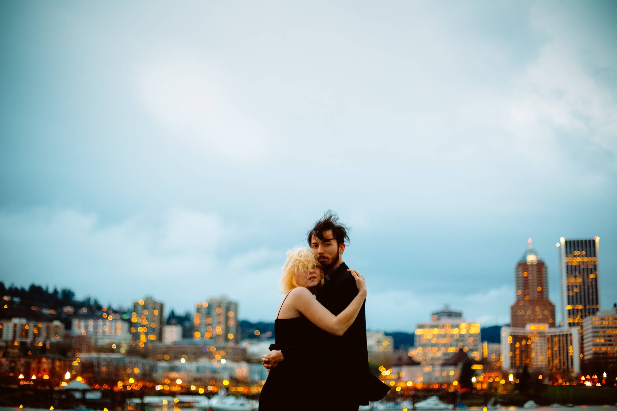A couple embraces in the foreground with a city skyline and glowing lights behind them at dusk. The sky is cloudy, and the atmosphere feels intimate and romantic. Jessica Hill Photography - Wedding Photographer - Oregon