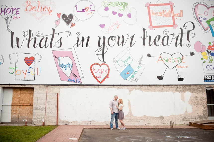 A couple stands in front of a mural reading "What's in your heart?" surrounded by colorful hearts and positive words on a white brick wall. Jessica Hill Photography - Wedding Photographer - Oregon
