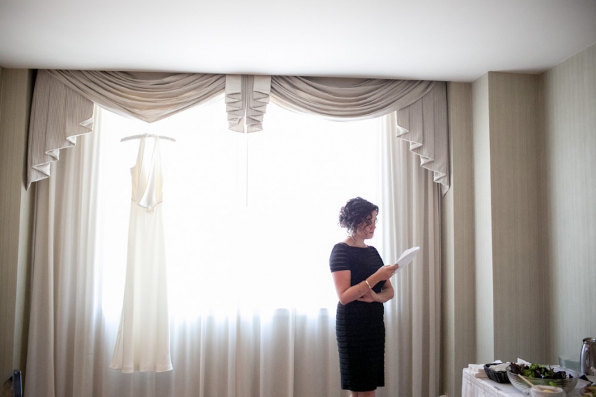 A woman in a black dress stands by a window reading a piece of paper, with a white dress hanging in the window beside her. Jessica Hill Photography - Wedding Photographer - Oregon