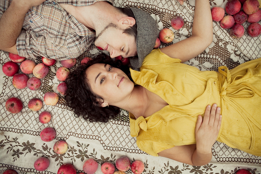 A man and a woman lie on a patterned blanket surrounded by apples. The woman in a yellow dress faces the camera, while the man in a hat rests beside her with eyes closed. Jessica Hill Photography - Wedding Photographer - Oregon