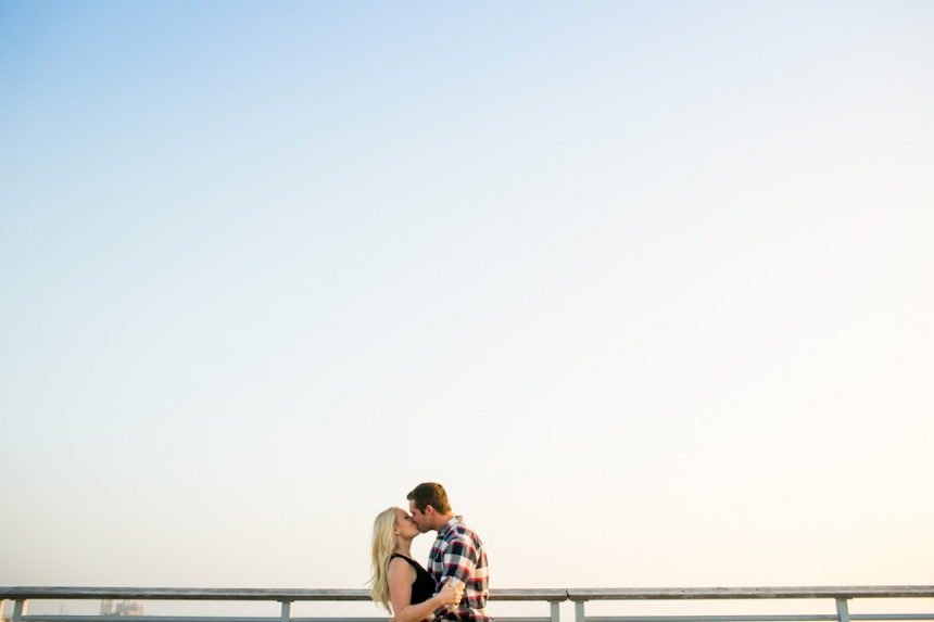 A couple stands close together and kisses by a railing under a clear sky, with ample empty space above them. Jessica Hill Photography - Wedding Photographer - Oregon