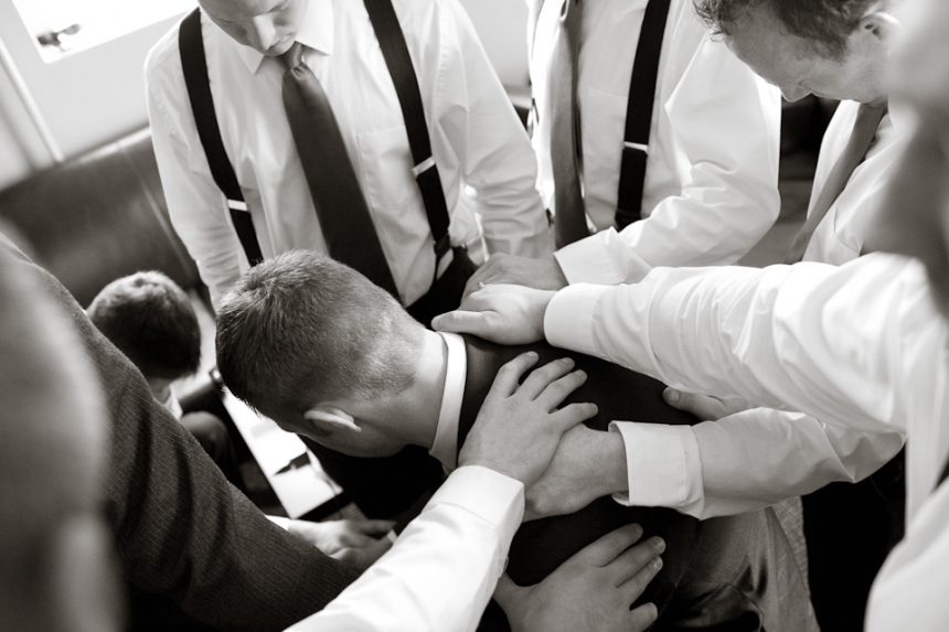 A group of men in dress shirts and suspenders stand around another man, placing their hands on his shoulders and back in a gesture of support. Jessica Hill Photography - Wedding Photographer - Oregon