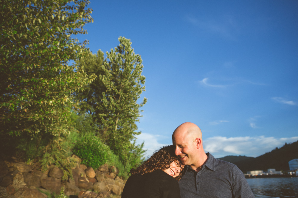 A woman rests her head on a man's shoulder as they stand outdoors near trees and water under a clear blue sky. Jessica Hill Photography - Wedding Photographer - Oregon