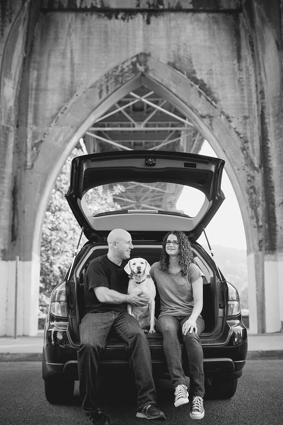 A man and woman sit in the open trunk of a car under a large bridge, with a dog sitting between them. Jessica Hill Photography - Wedding Photographer - Oregon