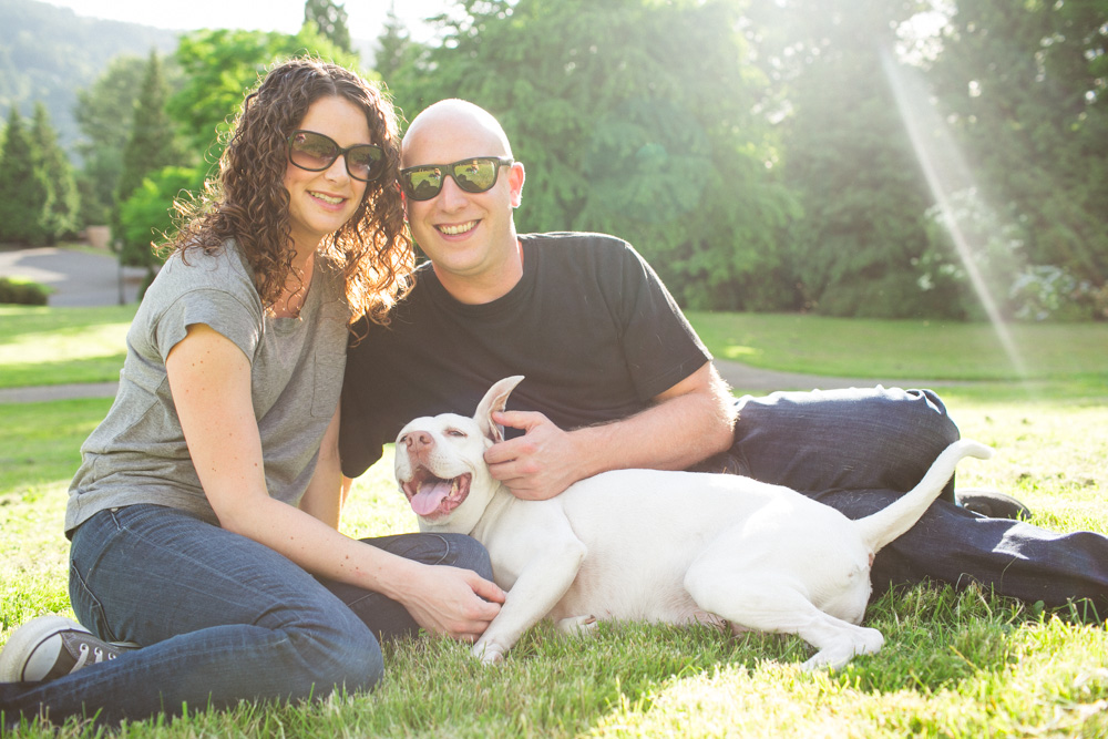 A man and woman sit on grass in a park, smiling at the camera with a white dog lying between them. Sunlight filters through trees in the background. Jessica Hill Photography - Wedding Photographer - Oregon