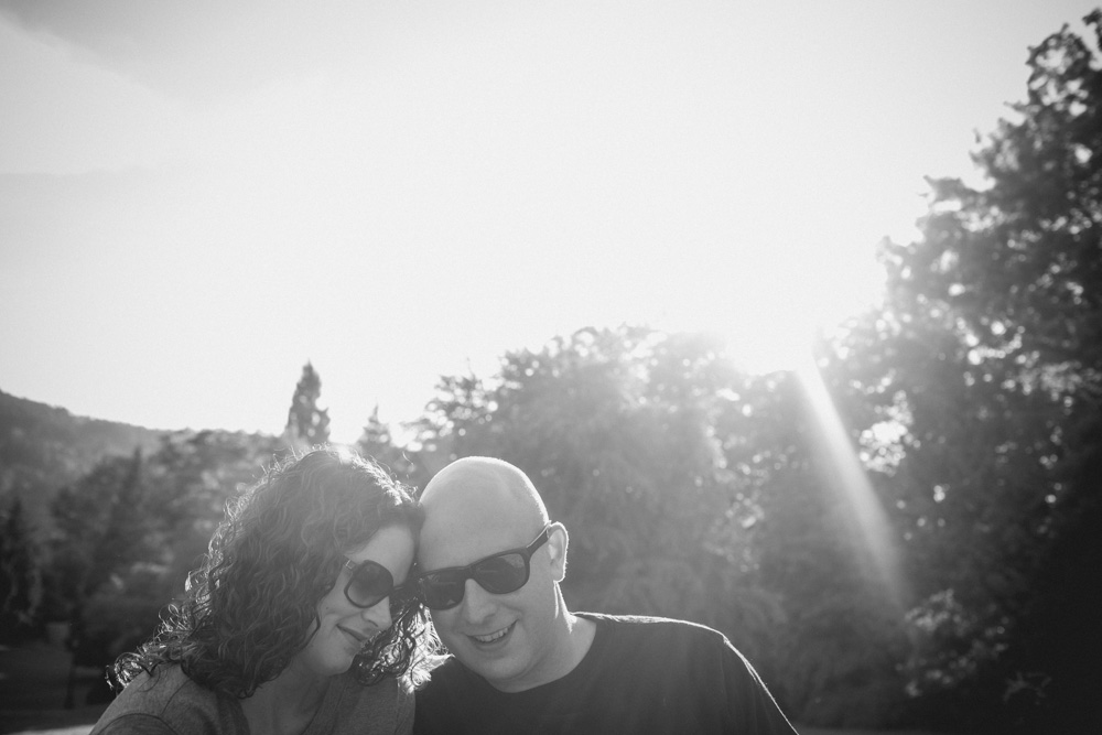 A man and a woman wearing sunglasses sit close together outdoors, with trees and bright sunlight in the background. Jessica Hill Photography - Wedding Photographer - Oregon