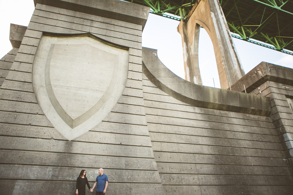 A man and woman hold hands at the base of a large concrete bridge support featuring a shield-shaped design. Jessica Hill Photography - Wedding Photographer - Oregon