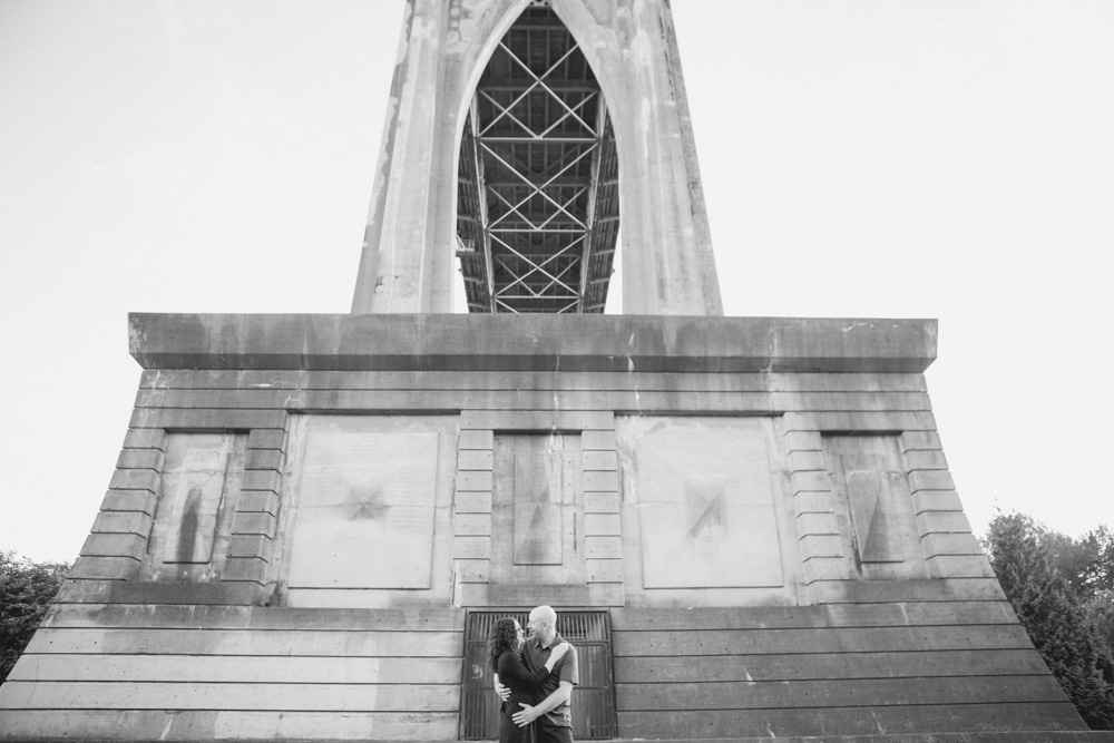 Two people hold hands while standing on a log beneath a large bridge at sunset, with trees and the sky in the background. Jessica Hill Photography - Wedding Photographer - Oregon