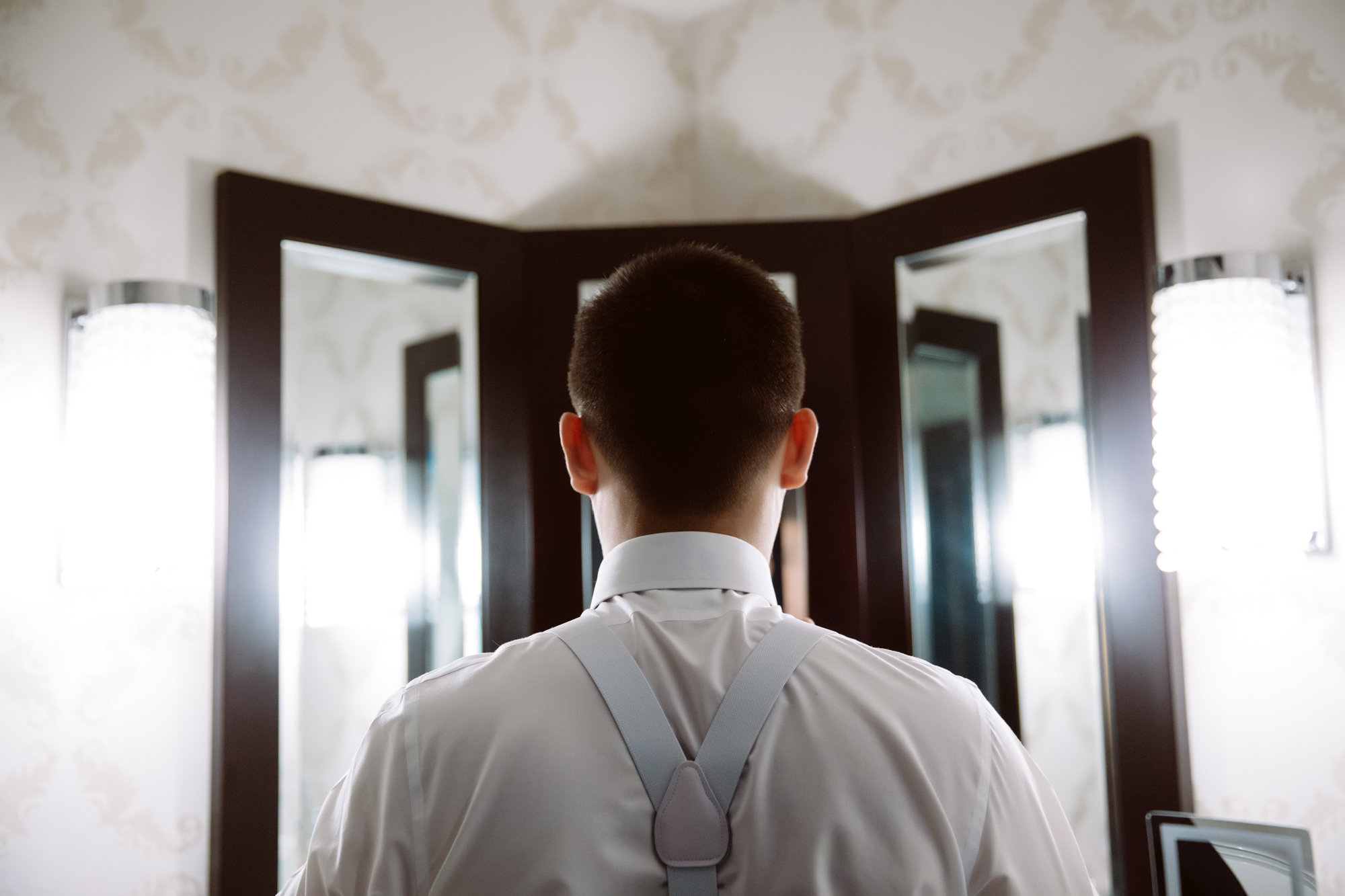 A man in a dress shirt and suspenders stands facing a three-panel mirror in a well-lit room, shown from behind. Jessica Hill Photography