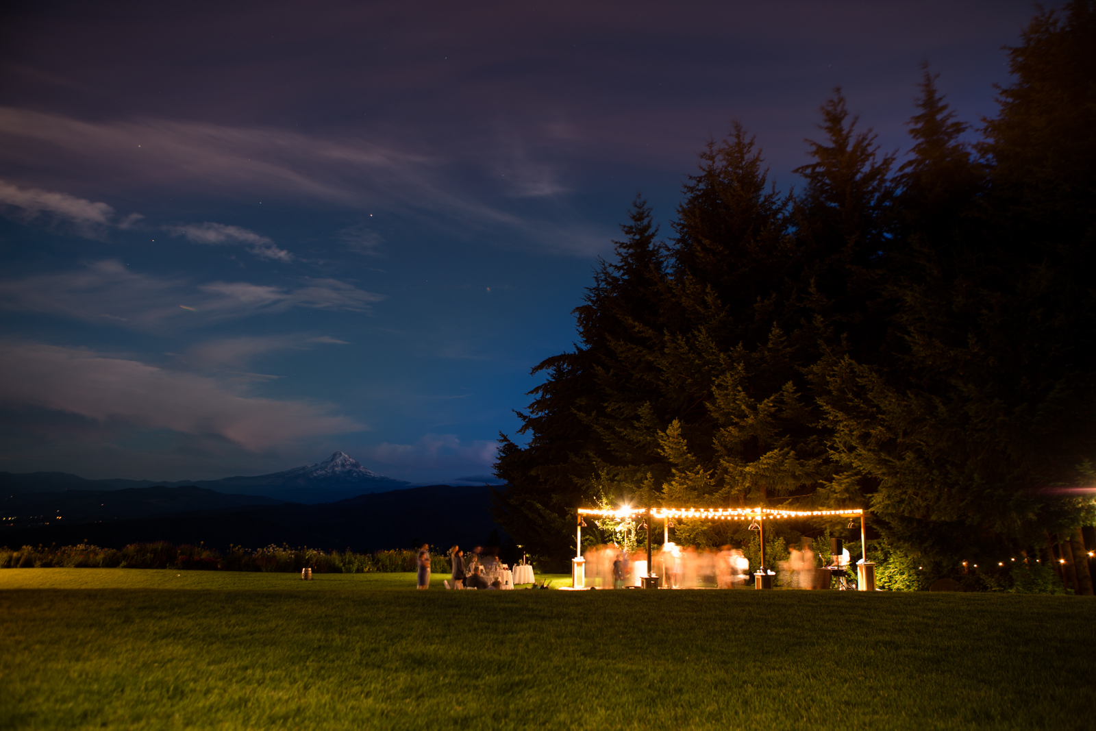 A couple stands close together, silhouetted against a cloudy evening sky with a city skyline and glowing building lights in the background. Jessica Hill Photography - Wedding Photographer - Oregon
