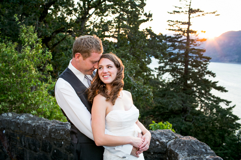 A couple dressed in wedding attire stands embracing near a stone wall, with trees and a body of water in the background at sunset. Jessica Hill Photography - Wedding Photographer - Oregon