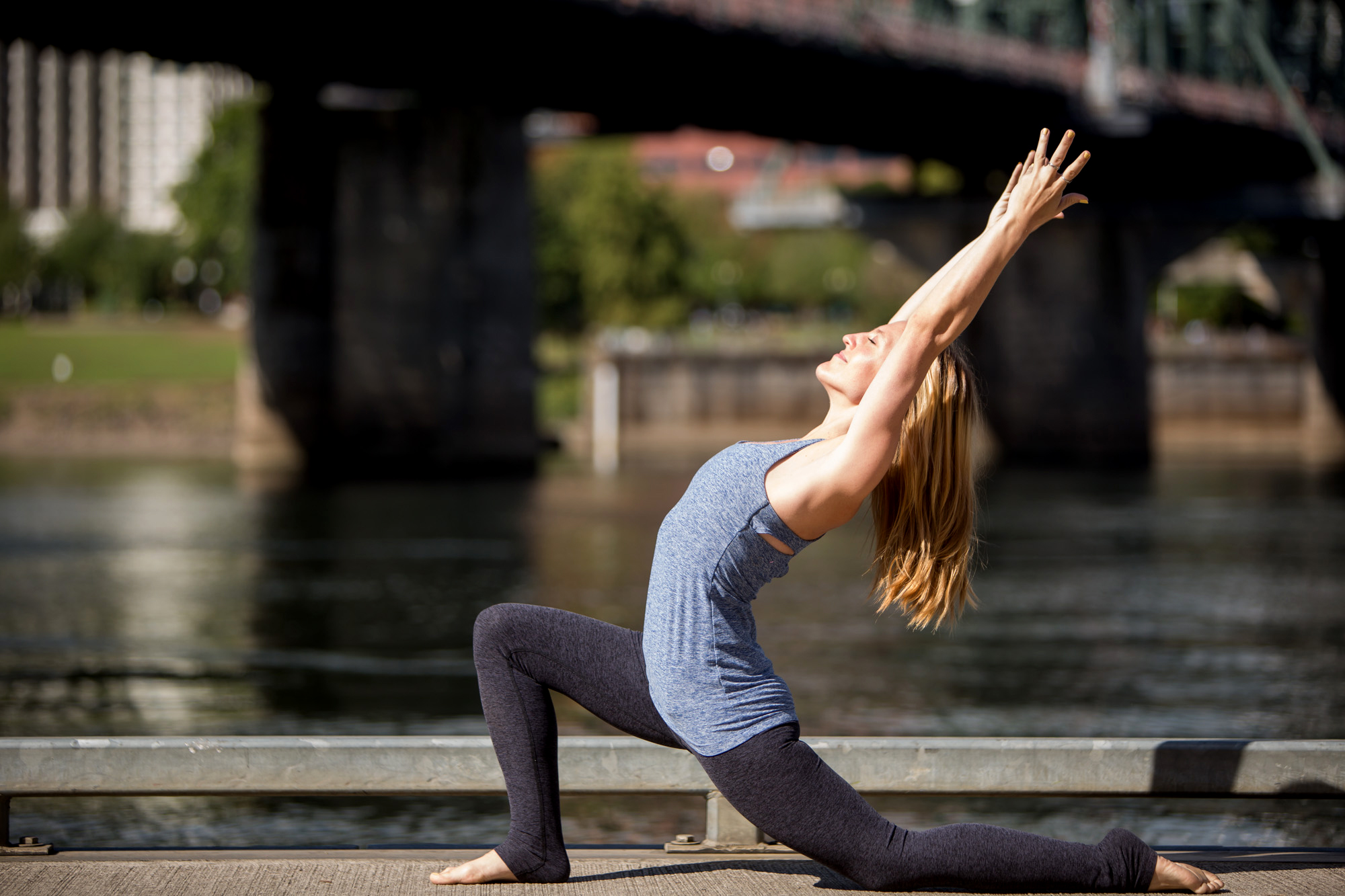 A woman practices yoga outdoors on a riverside pathway, performing a low lunge with her arms extended upward and back, beneath a bridge on a sunny day. Jessica Hill Photography - Wedding Photographer - Oregon