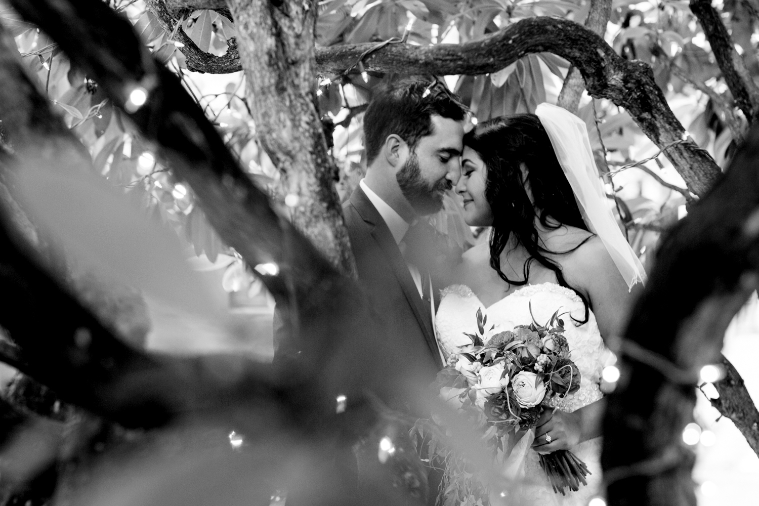 A bride and groom stand closely, touching foreheads and smiling, framed by tree branches with string lights. The bride holds a bouquet and wears a veil; the photo is in black and white. Jessica Hill Photography - Wedding Photographer - Oregon