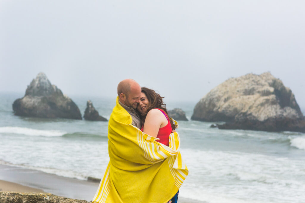 A couple wrapped in a yellow blanket smiles and hugs on a foggy beach, with large rocks and gentle waves in the background. Jessica Hill Photography - Wedding Photographer - Oregon