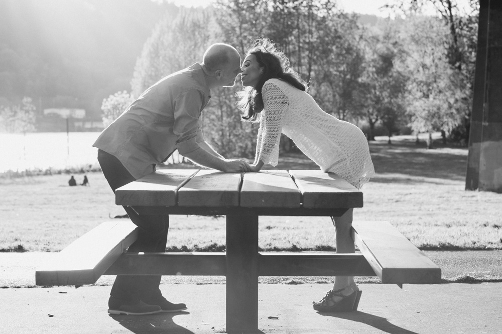 A man and woman lean toward each other over a picnic table outdoors, touching hands and faces close, with sunlight shining behind them. Jessica Hill Photography - Wedding Photographer - Oregon