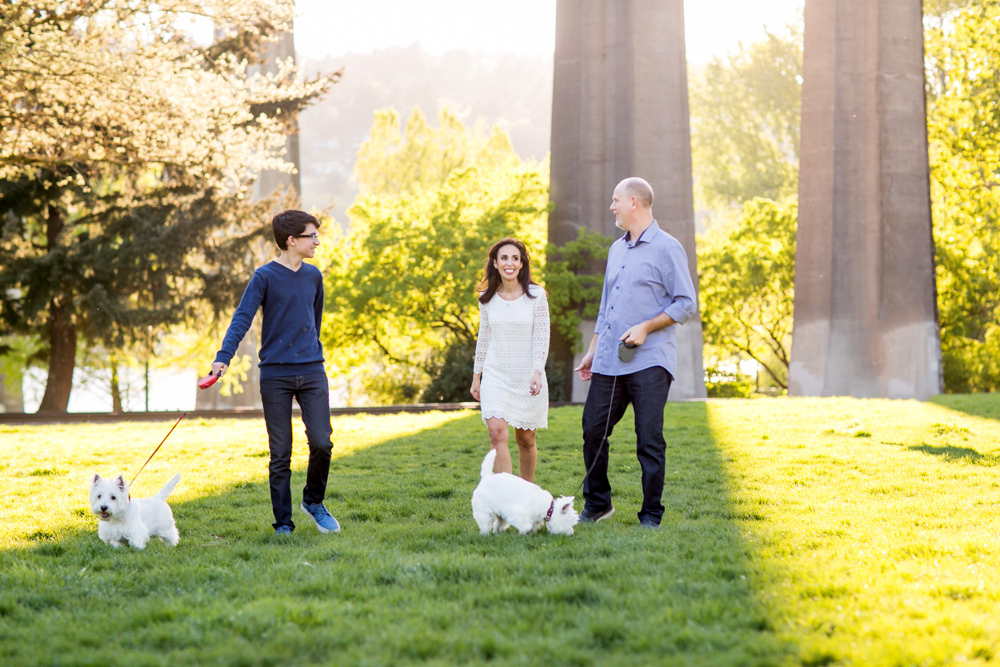 Three adults and two small white dogs walk on grass in a park with trees and large concrete pillars in the background. Jessica Hill Photography - Wedding Photographer - Oregon