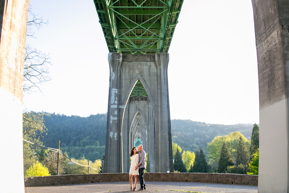 A couple stands close together under a large green bridge with concrete pillars, surrounded by trees and a distant hillside. Jessica Hill Photography - Wedding Photographer - Oregon