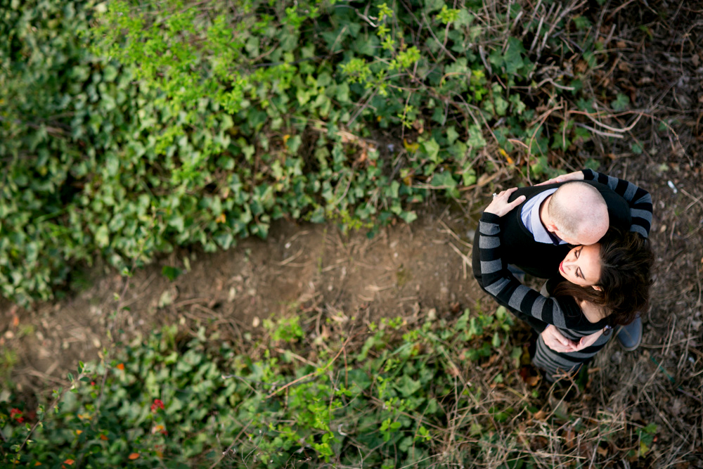A man and a woman embrace while standing on a narrow dirt path surrounded by green foliage, seen from above. Jessica Hill Photography - Wedding Photographer - Oregon