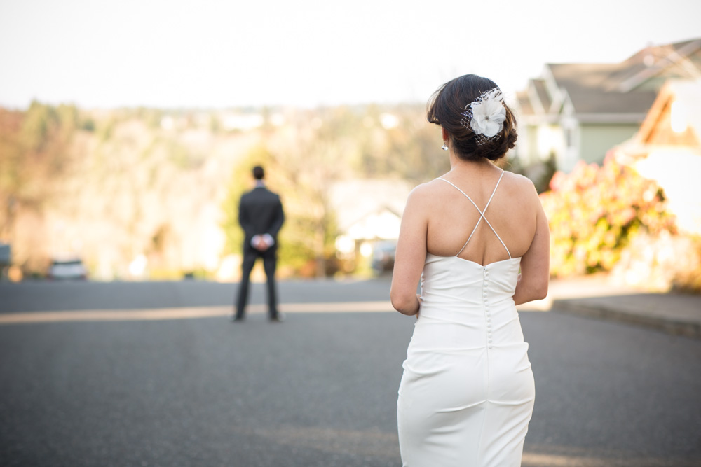 A woman in a white dress stands in the foreground, facing a man in a suit who is standing on a suburban street with his back to her. Jessica Hill Photography - Wedding Photographer - Oregon