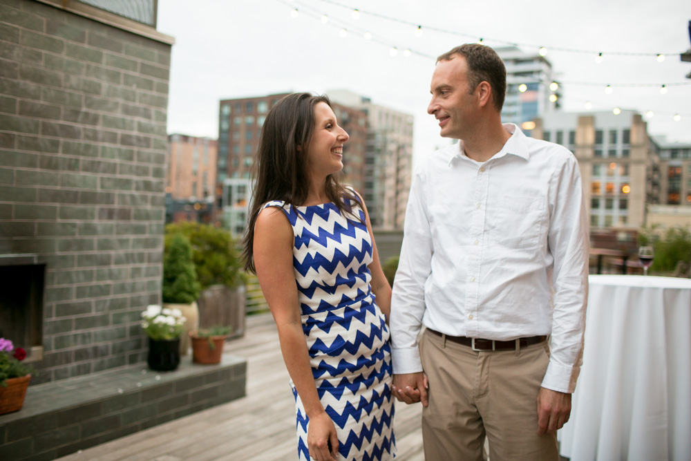 A woman in a blue-and-white zigzag dress and a man in a white shirt and khaki pants hold hands and smile at each other on a rooftop patio with city buildings in the background. Jessica Hill Photography - Wedding Photographer - Oregon
