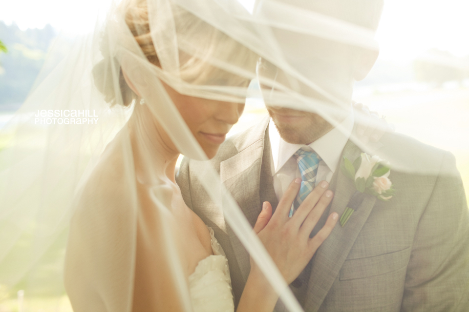 A bride and groom stand close together outdoors, partially covered by a sheer veil, with the bride’s hand resting on the groom’s chest. Jessica Hill Photography - Wedding Photographer - Oregon