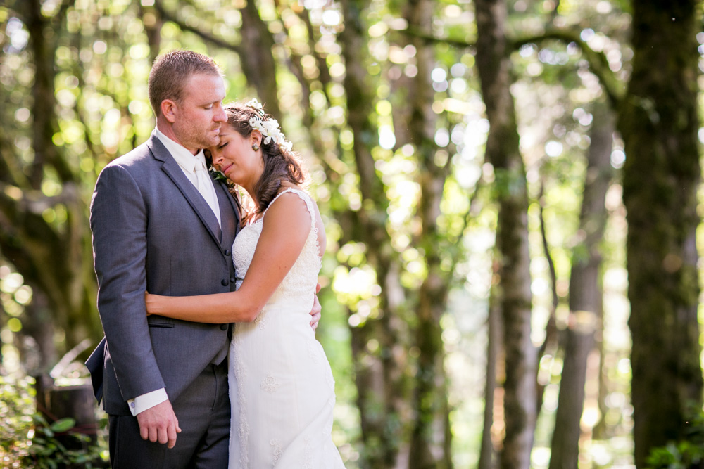 A bride and groom embrace in a sunlit forest. The groom, in a gray suit, gently kisses the bride’s forehead as she rests her head on his chest. Soft light filters through the trees around them. Jessica Hill Photography - Wedding Photographer - Oregon